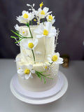 White handmade wafer paper daisies with yellow centres decorating a two-tier cake with delicate green stems.