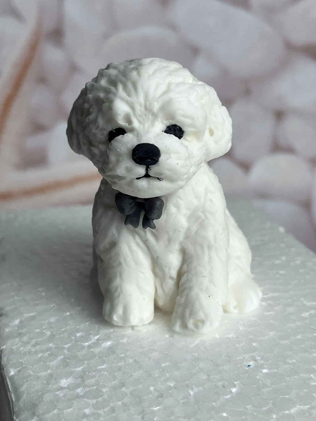 Close-up of a white edible fondant dog cake topper with a dark bow, black eyes and textured fur.